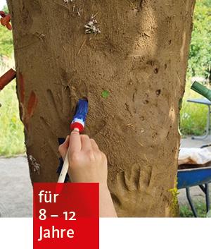 ein mit Lehm ummantelter Baumstamm. Im Lehm sind Handabdrücke von Kindern. Eine Kinderhand fährt mit einem Pinsel einen Handabdruck entlang