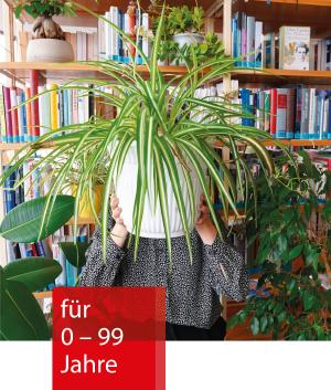 Vor einem Bücherregal steht eine Person, die sich eine Zimmerpflanze vor das Gesicht hält. Daneben stehen weitere Pflanzen