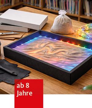 in einem Rahmen, der auf einem Tisch liegt, sind farbige Sandwellen. Um den Rahmen herum stehen ein Säckchen Sand, Bastelmaterial, Kartons