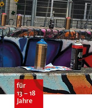 Farbspraydosen stehen vor einer besprühten Wand. Im Hintergrund ein hoher Metallzaun mit Tor