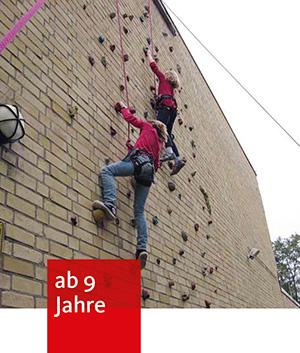 An einer Backsteinwand sind Klettergriffe befestigt. An diesen klettern zwei Kinder mit Kletterausrüstung nach oben.