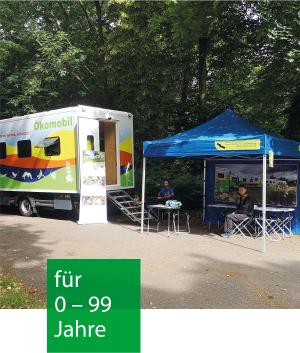 Auf einem asphaltierten Weg im Wald steht das Ökomobil mit offener Türe. Davor steht ein Pavillon mit Tischen und Stühlen