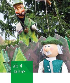 Zwei Marionetten vor einer gebauten Kulisse und Nadelbäumen.