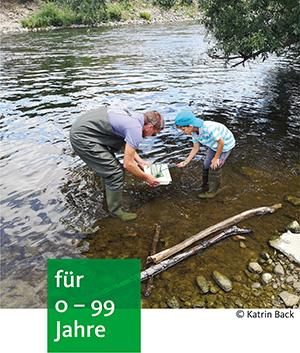 ein Erwachsener und ein Kind stehen in Gummistiefel am Flussrand. Sie sind gebückt und schauen in eine Schale