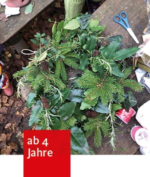 Ein grüner Kranz aus Tannenzweigen, Efeu, und anderen Blättern. Daneben liegen Bastelmaterialien wie Schere, rotes Band, Schnur